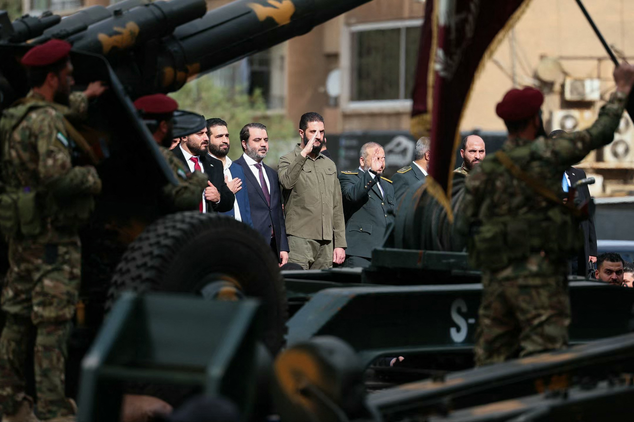 This handout photograph released by the official Syrian Arab News Agency (SANA) on December 8, 2025, shows Syria's President Ahmed al-Sharaa (CR) saluting and Syrian Minister of Foreign Affairs and Expatriates Asaad Al-Shaibani (CL) observing the military parade in Damascus during the anniversary of the ousting of longtime ruler Bashar al-Assad.