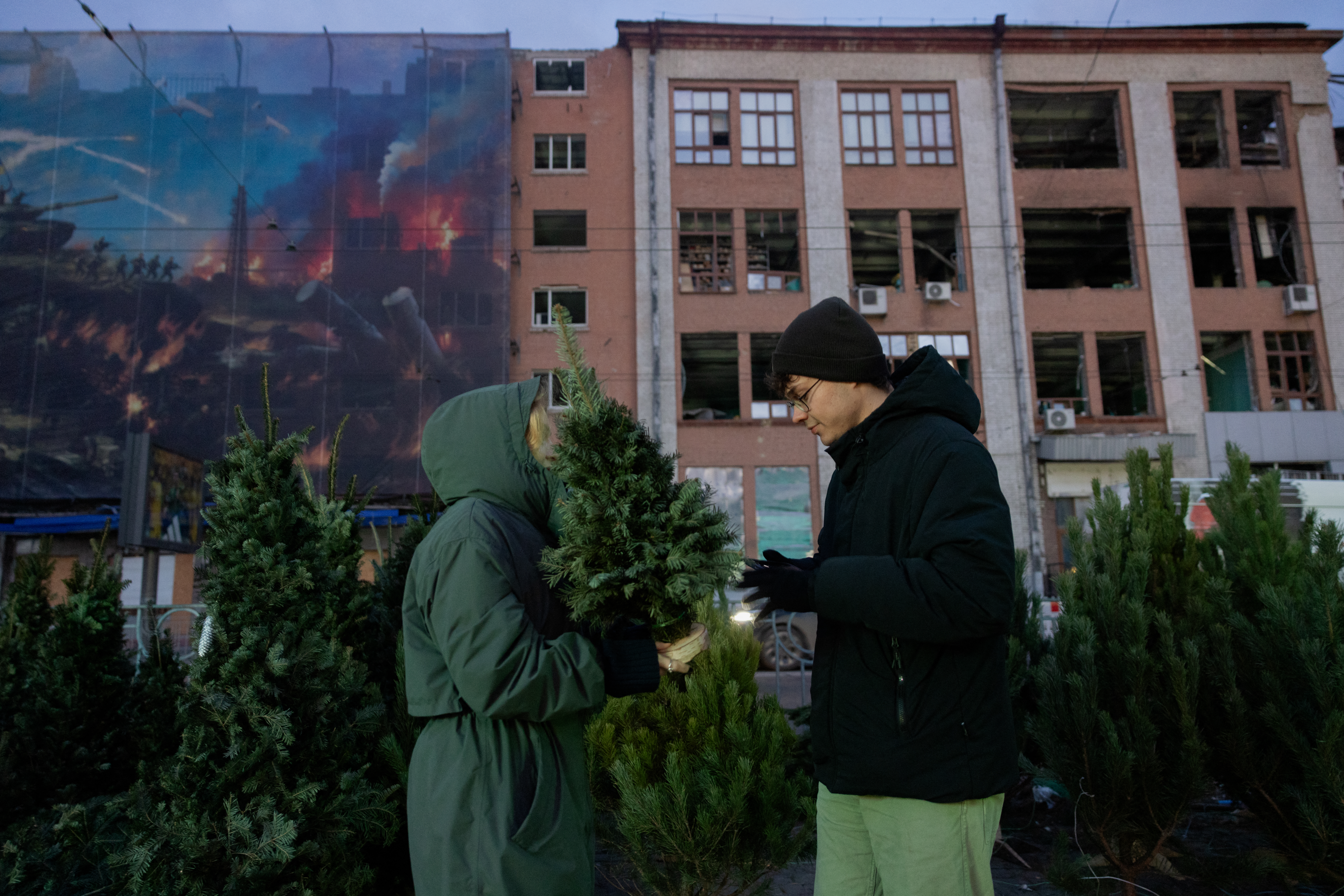 A woman (L) holds a Christmas tree at a street market near damaged buildings.