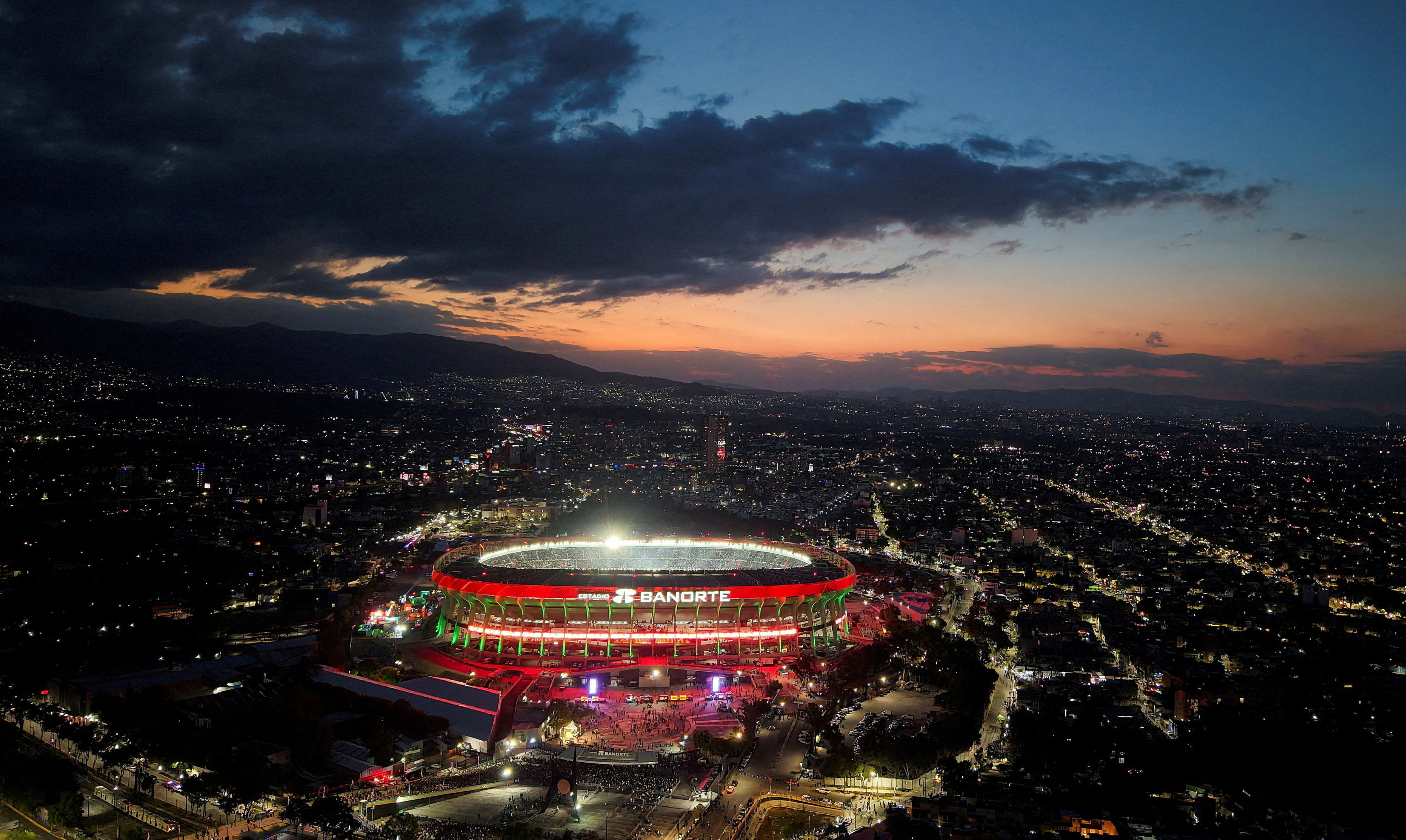 Azteca Stadium lit up at sunset with a dark city behind and clouds overhead
