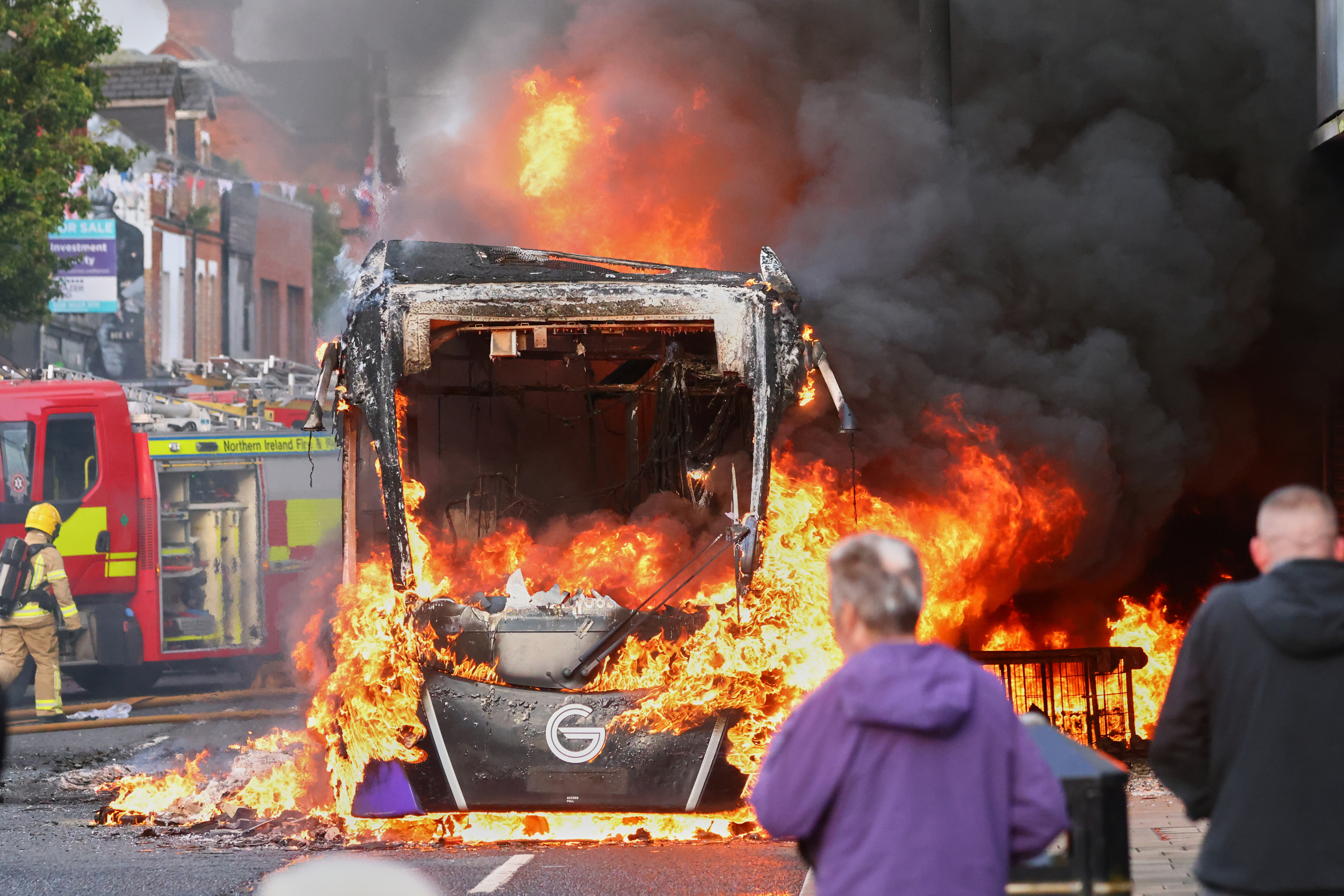 People watch as firemen arrive to put out vehicle that was set alight during a protest in East Belfast