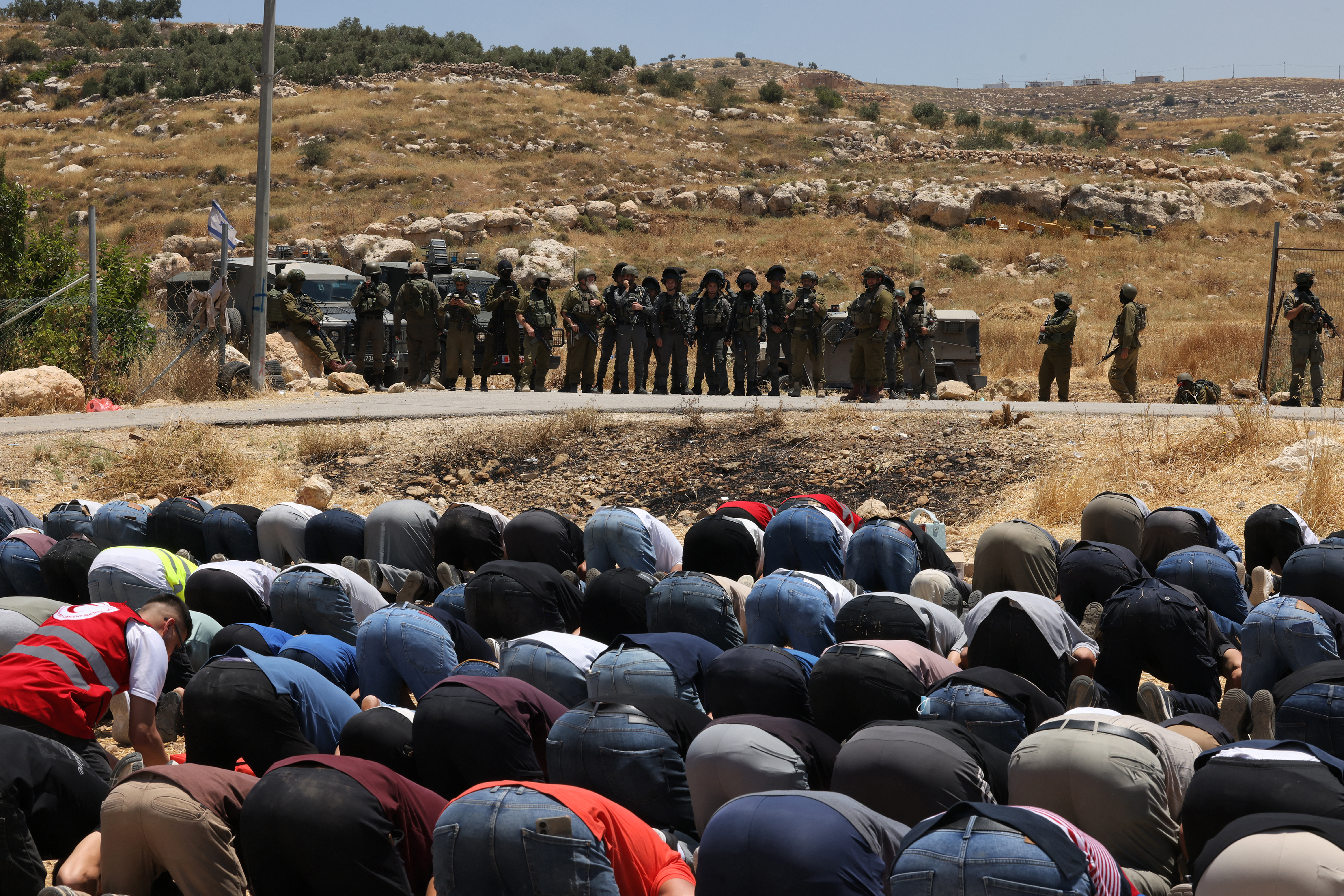 Israeli security forces stand guard as Palestinian men, who had gathered to protest threats of confiscation of their land, perform the Muslim Friday noon prayers at a field in the Palestinian village of Idna, west of Hebron in the occupied West Bank, on June 12, 2026.