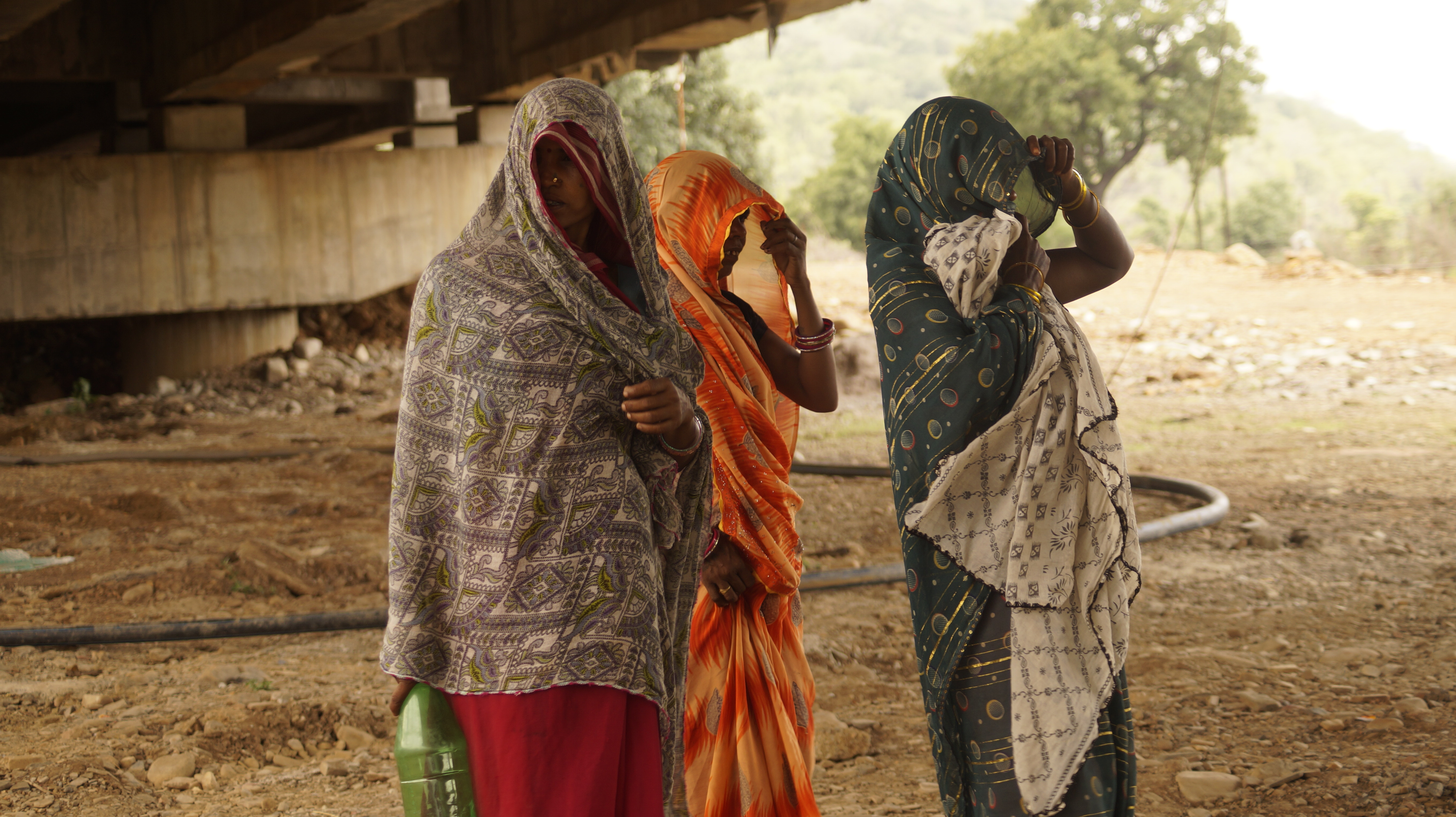 Three women stand together, covering their faces with their sarees.
