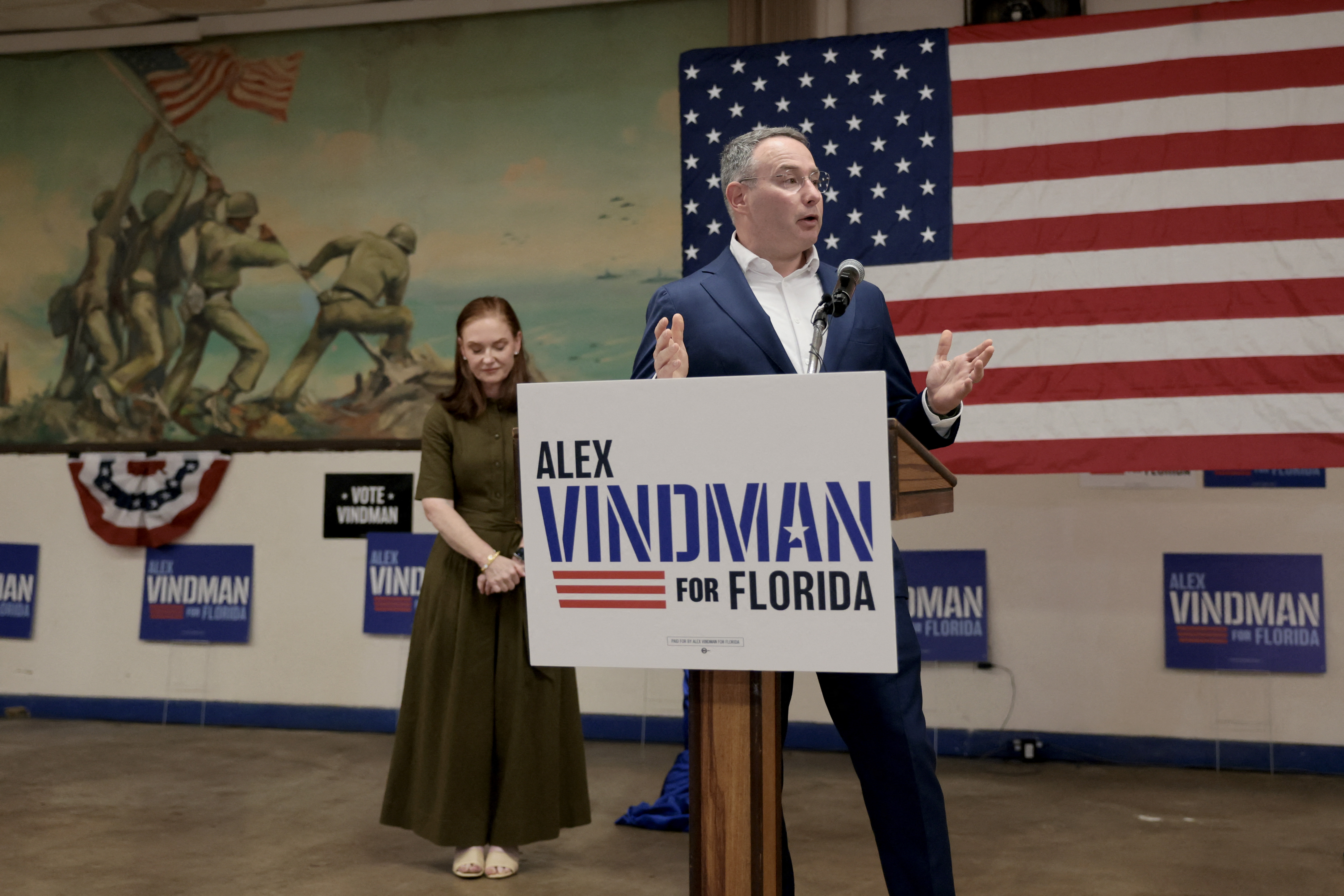 A man in a suit stands next to a campaign poster